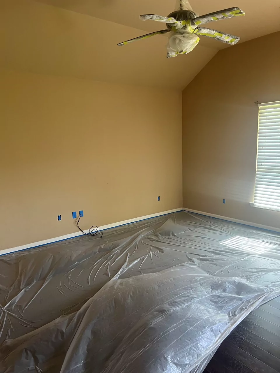A bedroom prepared for painting: the floor fully covered in plastic sheeting, the ceiling fan wrapped, and the outlet openings taped off along a freshly cut-in wall.