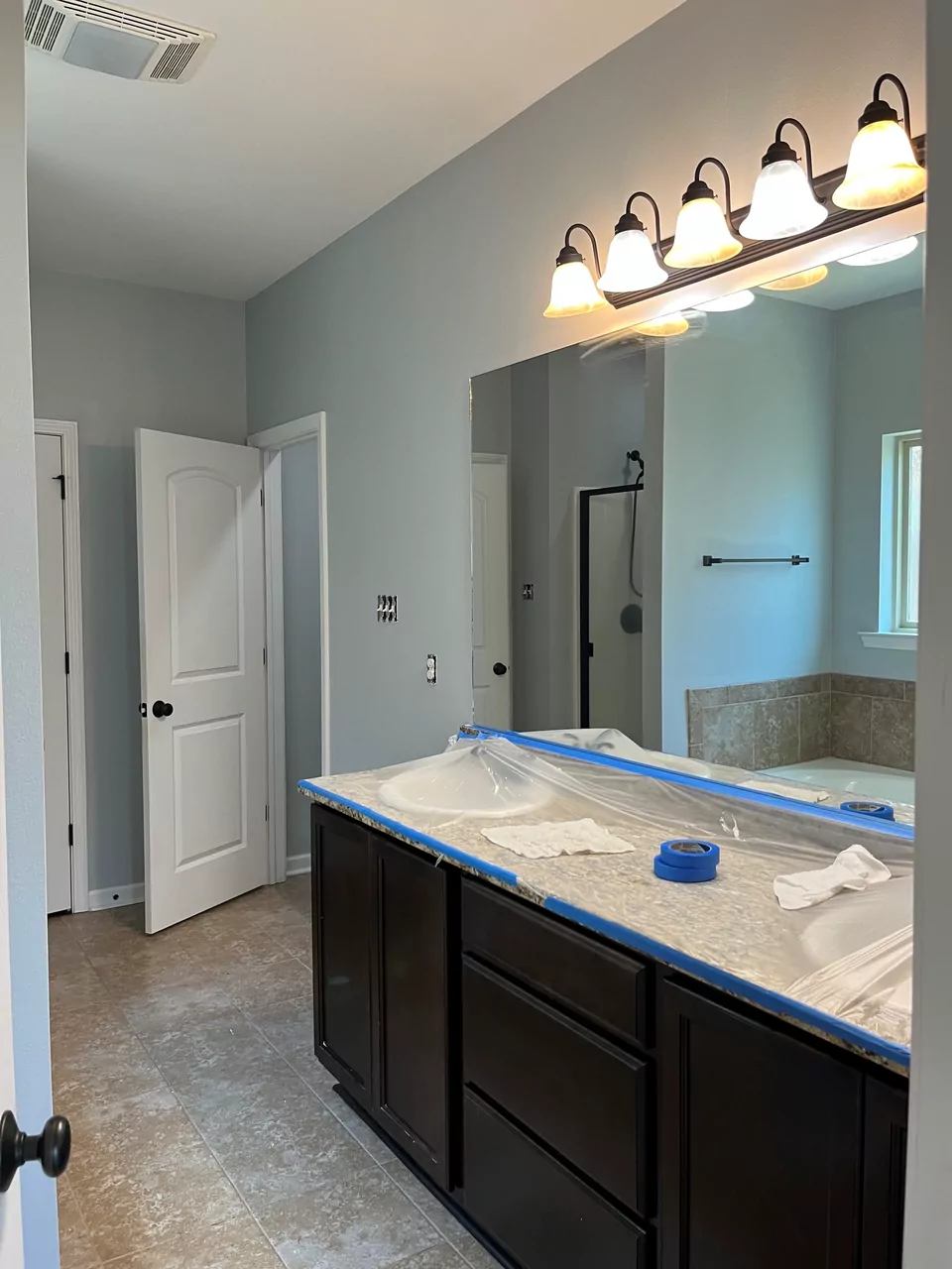 A bathroom mid-repaint in soft grey, with the vanity top masked in plastic and blue tape and the mirror and light bar protected.